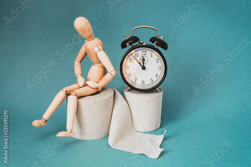 Wooden figure sit on a roll of toilet paper near alarm clock. Concept of the problem with digestion.