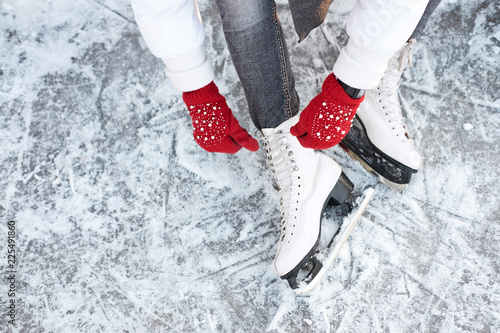 Girl tying shoelaces on ice skates before skating on the ice rink, hands in red knitted gloves.