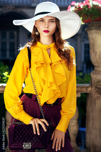 Outdoor fashion portrait of young beautiful woman wearing stylish white hat, yellow blouse with frills, violet trousers, holding velvet quilted bag, posing in street, near old european architecture