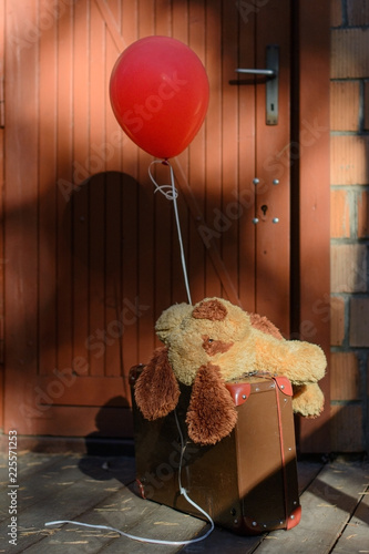 Pluszowy piesek leżący na walizce z czerwonym balonem przed drzwiami domu
