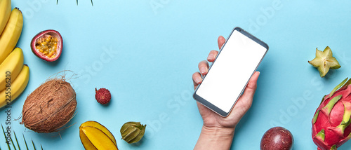 Coconut, lychee, pineapple set of tropical fruits. The girl's hand is holding the phone on a blue background with a copy of the space. The concept of modern online shopping. Flat lay