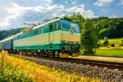 A green electric locomotive passing the Czech countryside. A train running through the green valley. Rail transport in the Czech Republic. A sunny day on the railroad. 