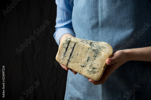 Woman in apron holding piece of blue cheese Gorgonzola on dark background