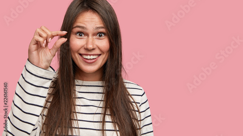 Cropped shot of glad dark haired young woman shows very tiny, gestures with hand, dressed in casual clothes, poses over pink background with free space for your promotion. Look, its too small