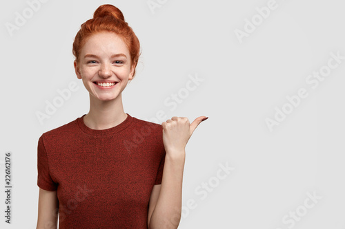 Advertisement concept. Cheerful foxy girl with tender toothy smile, freckled skin indicates with thumb aside, shows something over blank space, dressed in casual outfit, isolated on white background.