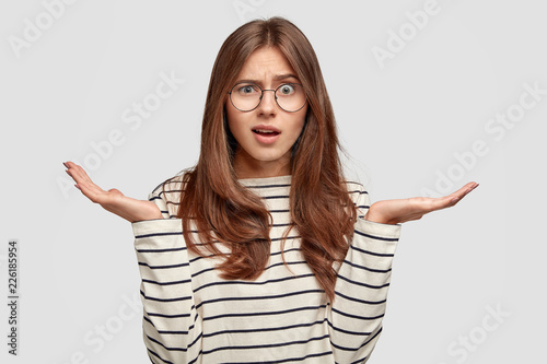 Questioned indignant pleasant looking brunette woman speads hands in hesitant gesture, wears striped casual sweater, looses something, forgets about some details, has frustrated facial expression