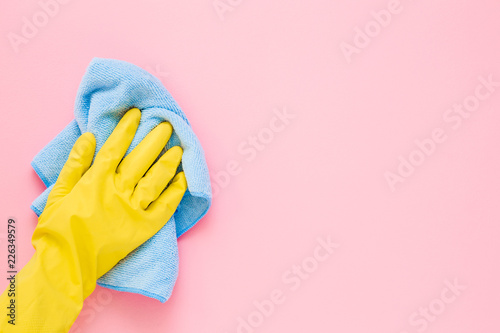 Employee hand in yellow rubber protective glove wiping pastel pink wall from dust with blue dry rag. General or regular cleanup. Commercial cleaning company. Copy space. Empty place for text or logo.