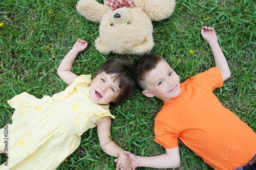 Top view of kids lying on the grass at park having fun. Little girl and boy relax with smiling. Teddy bear toy together