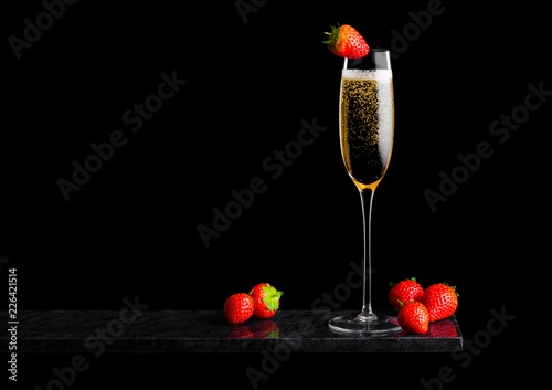 Elegant glass of yellow champagne with strawberry on top and fresh berries on black marble board on black background. Space for text