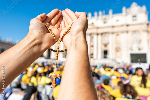 Macro closeup of hands holding wooden handmade Italian cross Catholic Assisi rosary with bokeh background of Vatican church St Peter's Square Basilica during mass on sunny day