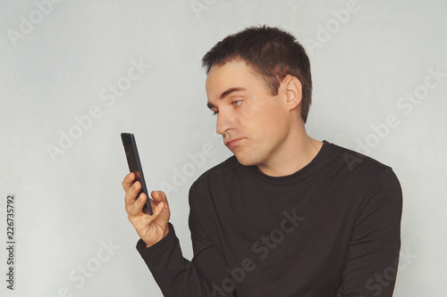 a young guy looks at the screen of a black smartphone with a dismissive emotion on his face. uninteresting correspondence. boring pastime.