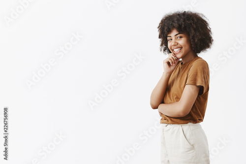 Intrigued flirty feminine dark-skinned female student with afro hairstyle in brown t-shirt standing in profile, turning at camera with cute smile holding hand on chin gazing with interest and desire