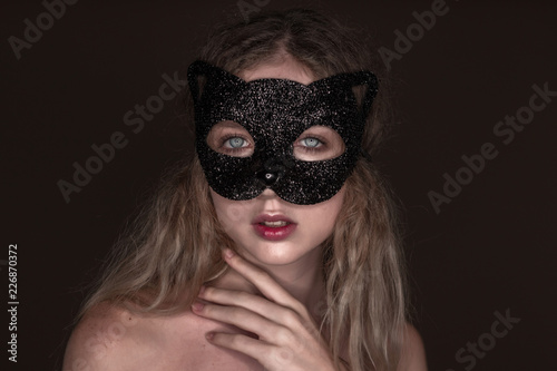 Beautiful young tender woman posing isolated over dark beige chocolate brown background wall wearing carnival cat masquerade mask looking camera.