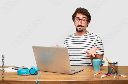 young bearded graphic designer with a laptop with a quizzical and confused look, doubting between different choices or possibilities with a wondering expression, comparing and posing a question.