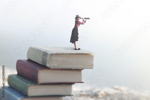 miniature woman looks at the infinity with the spyglass on a scale of books