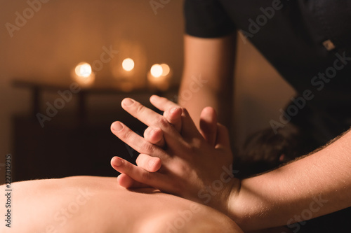 Men's hands make a therapeutic neck massage for a girl lying on a massage couch in a massage spa with dark lighting. Close-up. Dark Key