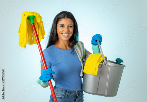 Young attractive woman holding cleaning tools and products in bucket isolated on blue background