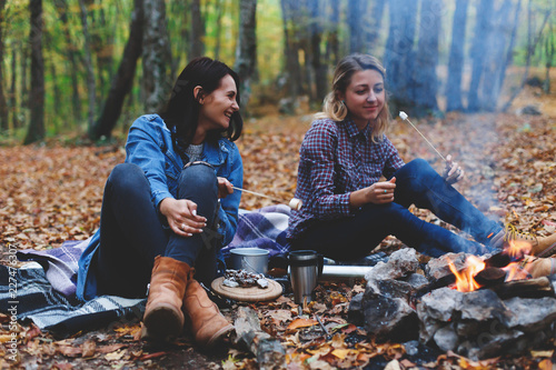 Two young girls girlfriends roasting sweet marshmallow on a fire in the evening in the autumn forest.