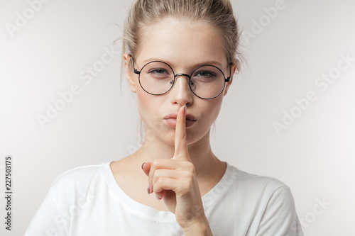 Cute mysterious girl wearing eyeglasses in casual outfit showing silence sign holding forefinger on lips, or having a big secret isolated on white background