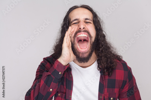 Portrait of roar handsome man with beard and black long curly hair in casual style, checkered red shirt standing and screaming with big open mouth. indoor studio shot, isolated on grey background.