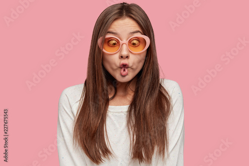 Funny playful Caucasian woman makes fish lips, has crazy look, tries to amuse depressed friend, feels bored, wears trendy shades and white sweater, isolated over pink background. Facial expressions