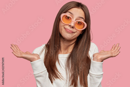 Indoor shot of clueless dark haired pretty young woman with doubtful expression, spreads palms with hesitation, wears casual white jacket and sunglasses, isolated over pink background. What to do?