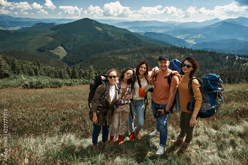 Top view full length portrait of four women and man standing and hugging. They hiking together through marvelous highland while carrying rucksacks and guy holding camera