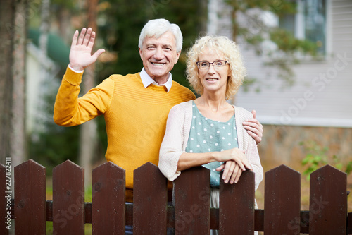 Hospitable mature couple standing by fence while friendly man waving hand to their neighbours