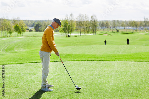 Senior man in casualwear standing in the middle of green field and going to hit golf ball