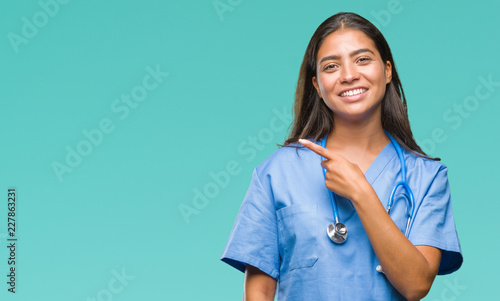 Young arab doctor surgeon woman over isolated background cheerful with a smile of face pointing with hand and finger up to the side with happy and natural expression on face looking at the camera.