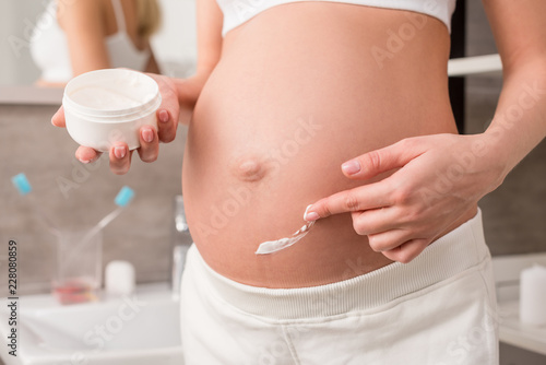 cropped shot of pregnant woman applying cream on tummy to avoid stretch marks in bathroom