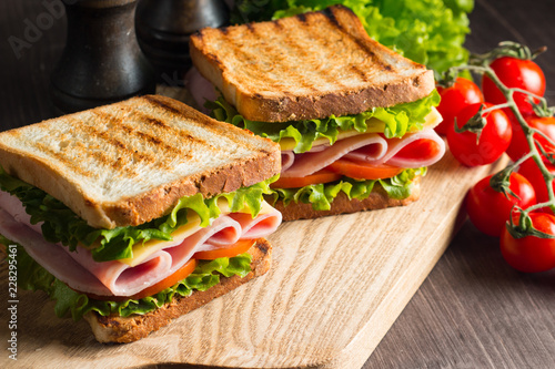 Close-up of two sandwiches with bacon, salami, prosciutto and fresh vegetables on rustic wooden cutting board. Club sandwich concept.