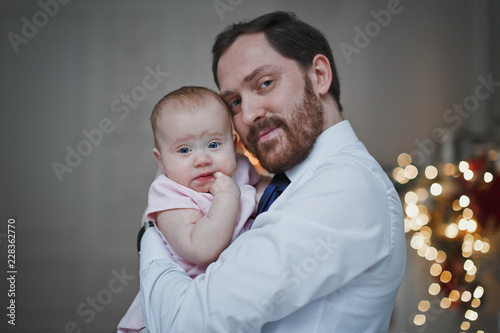 Happy father hugging his baby daughter for the Christmas tree lights in the background. Family, fatherhood and holidays concept