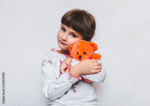 A little girl is hugging a soft orange teddy bear on a white background. Favorite toys of children. Orphanage.