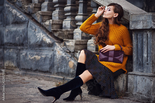Outdoor full body fashion portrait of young beautiful woman wearing color sunglasses, stylish orange sweater, polka dot skirt, trendy sock boots, holding small purple bag, posing in street. Copy space