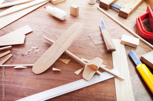 Making model airplane from wood. Wooden air plane handcrafted with balsa wood, on work table by the window. Airplane, cutter knife, balsa wood material and glue on table.