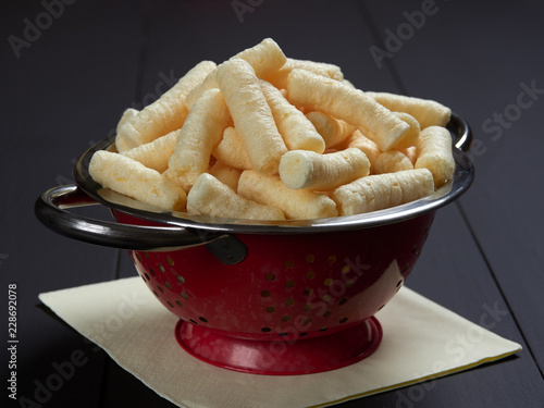 Crunchy, salty corn puffs snacks, also known in Romanian as pufuleti, in a red strainer, on dark brown background