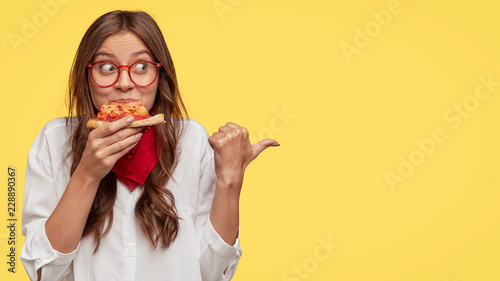 Photo of attractive woman eats slice of pizza, points aside with thumb, dressed in fashionable clothes, shows where pizzeria is, isolated over yellow background. Pretty girl has snack with fastfood