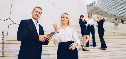 Boss keeping tablet and speaking with blonde secretary on stairs with biz partners in background. Concept of decision making and successful ideas. Business persons talking and waiting for conference.