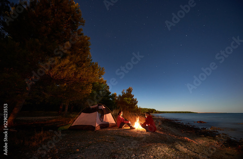 Night camping on sea shore. Happy young man and woman hikers relaxing in front of tent at campfire under evening sky on clear blue water and green forest background. Active lifestyle concept.