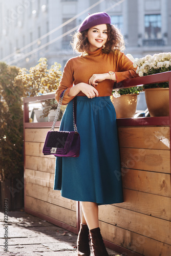 Outdoor full body fashion portrait of young beautiful happy smiling woman wearing stylish beret, orange turtleneck, midi green blue skirt, ankle boots, holding quilted purple bag, posing in street 