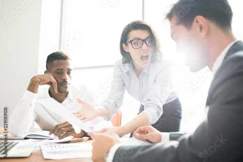 Furious displeased lady boss in glasses pointing at report and shouting at subordinate at staff meeting, business problems