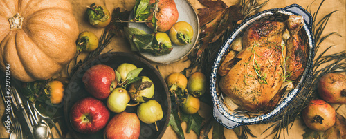Thanksgiving dinner table. Flat-lay of roasted chicken or turkey, fruit, pumpkin, cutlery, leaves over yellow table runner on grey concrete background, top view, wide composition