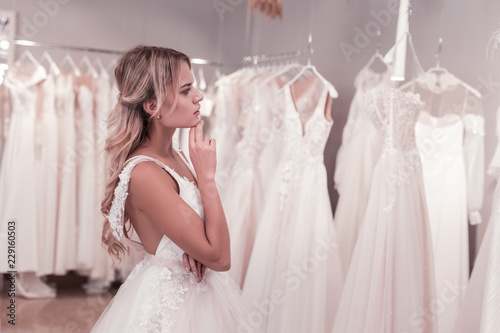 Which one is best. Pleasant thoughtful bride looking at the dresses while thinking which one to choose