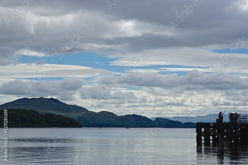Luss, Scotland: People taking selfies on a dock on Loch Lomond, with mountains on the far side of the lake. Loch Lomond is part of the Loch Lomond and The Trossachs National Park, established in 2002.