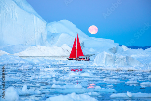 Little red sailboat cruising among floating icebergs in Disko Bay glacier during midnight sun season of polar summer. Ilulissat, Greenland.