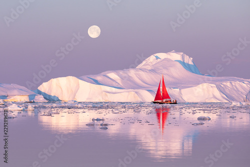 Little red sailboat cruising among floating icebergs in Disko Bay glacier during midnight sun season of polar summer. Ilulissat, Greenland.