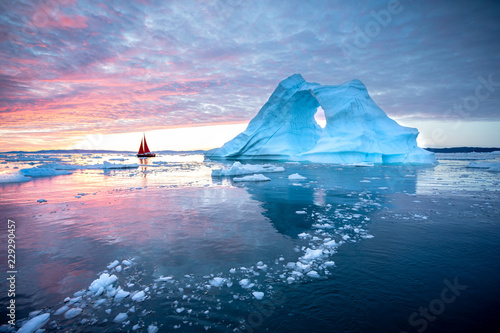 Little red sailboat cruising among floating icebergs in Disko Bay glacier during midnight sun season of polar summer. Ilulissat, Greenland.