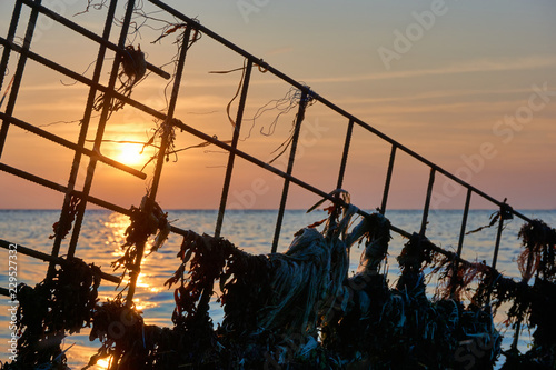 Silhouette of a metal structure with plastic waste and seaweed in front of the sea and sunset 