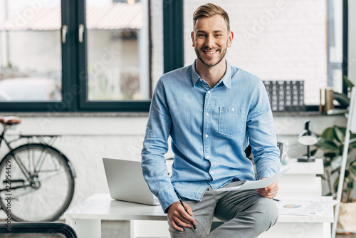 handsome young businessman holding papers and smiling at camera in office